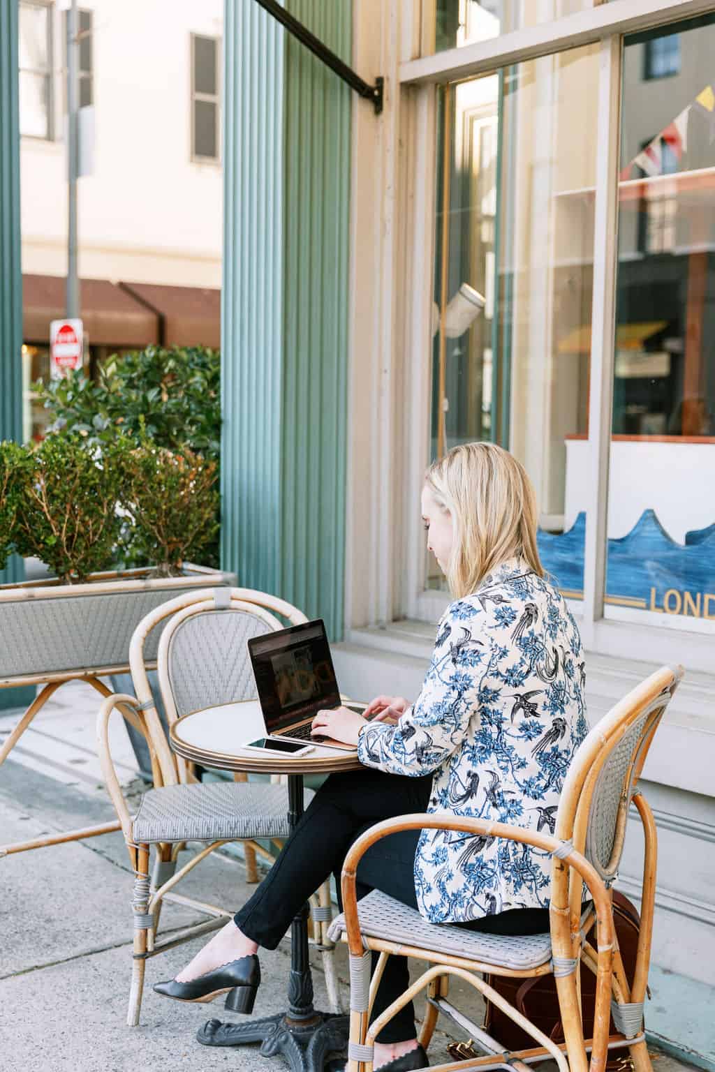 Blonde website designer sitting at cafe table outdoors working on her laptop in blue patterned blazer