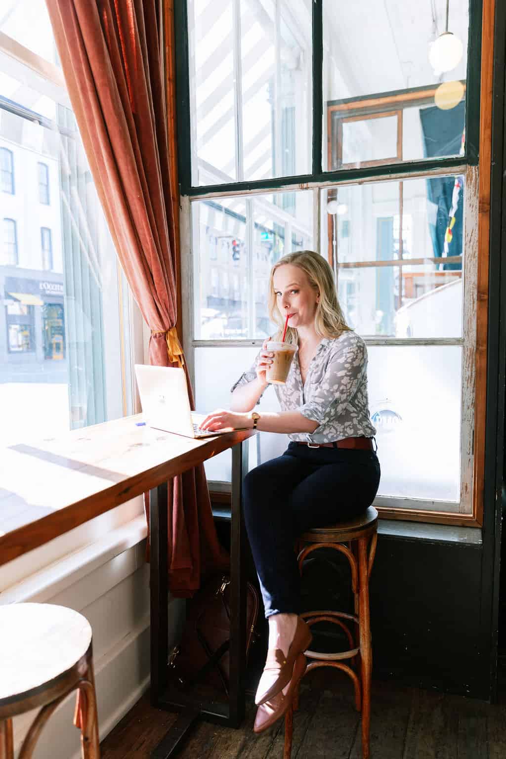 Digital Grace Design owner, Sarah Blodgett, sits on wooden stool in a cafe at her laptop drinking iced coffee